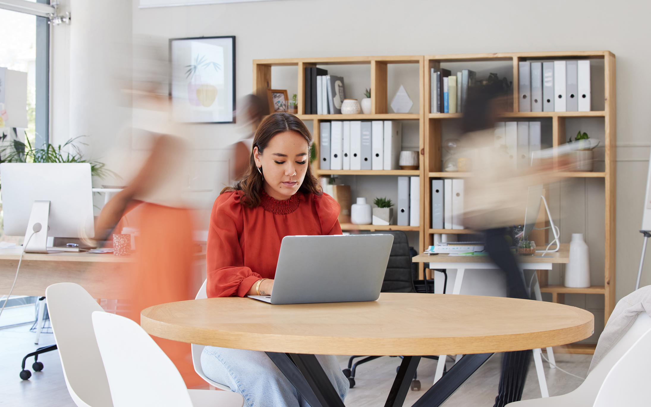 Serious asian businesswoman browsing internet on laptop in busy office. Focused ethnic professional working while colleagues rush in motion blur. Entrepreneur and creative designer using technology